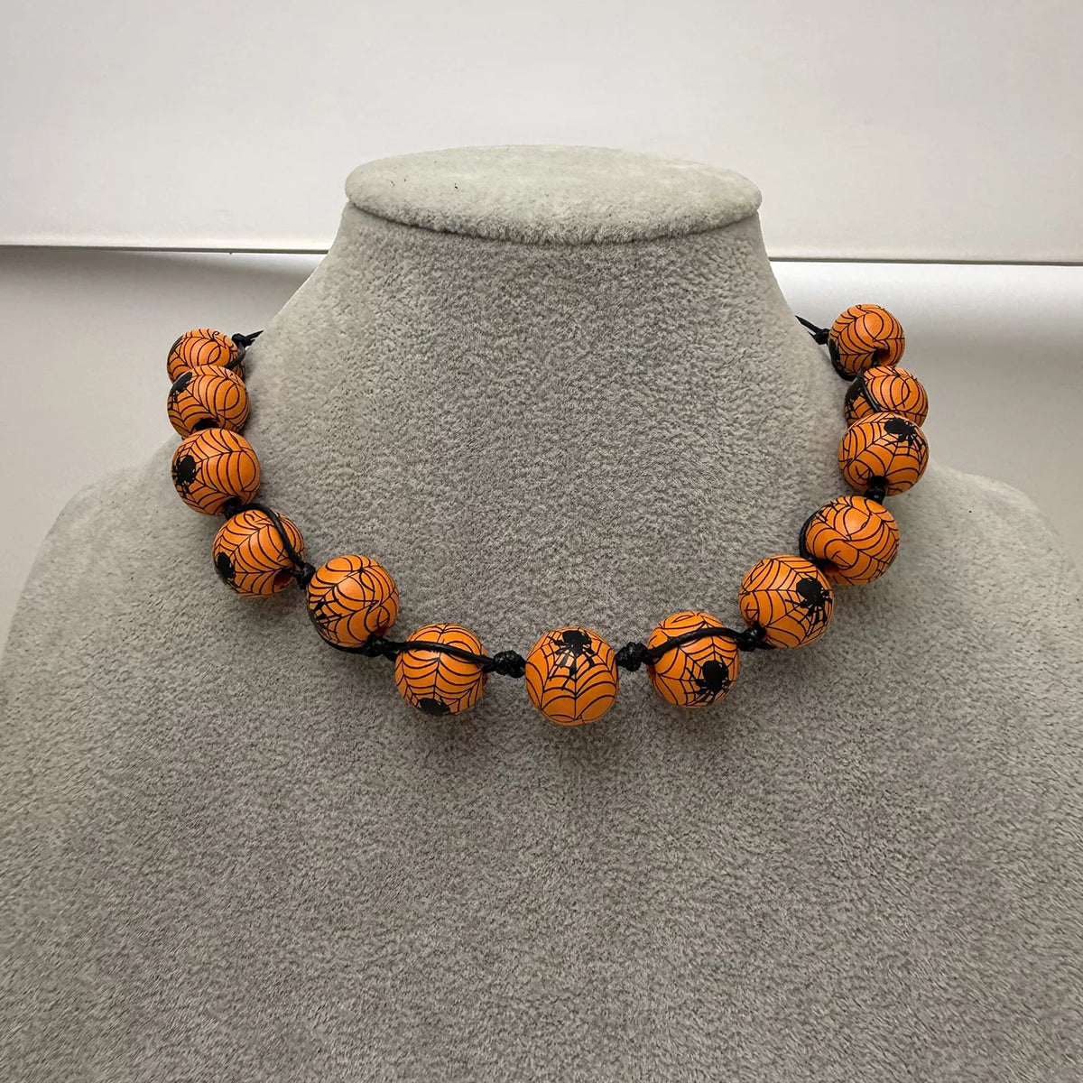An orange beaded necklace with black spiderweb and spider designs, displayed on a gray fabric jewelry bust.
