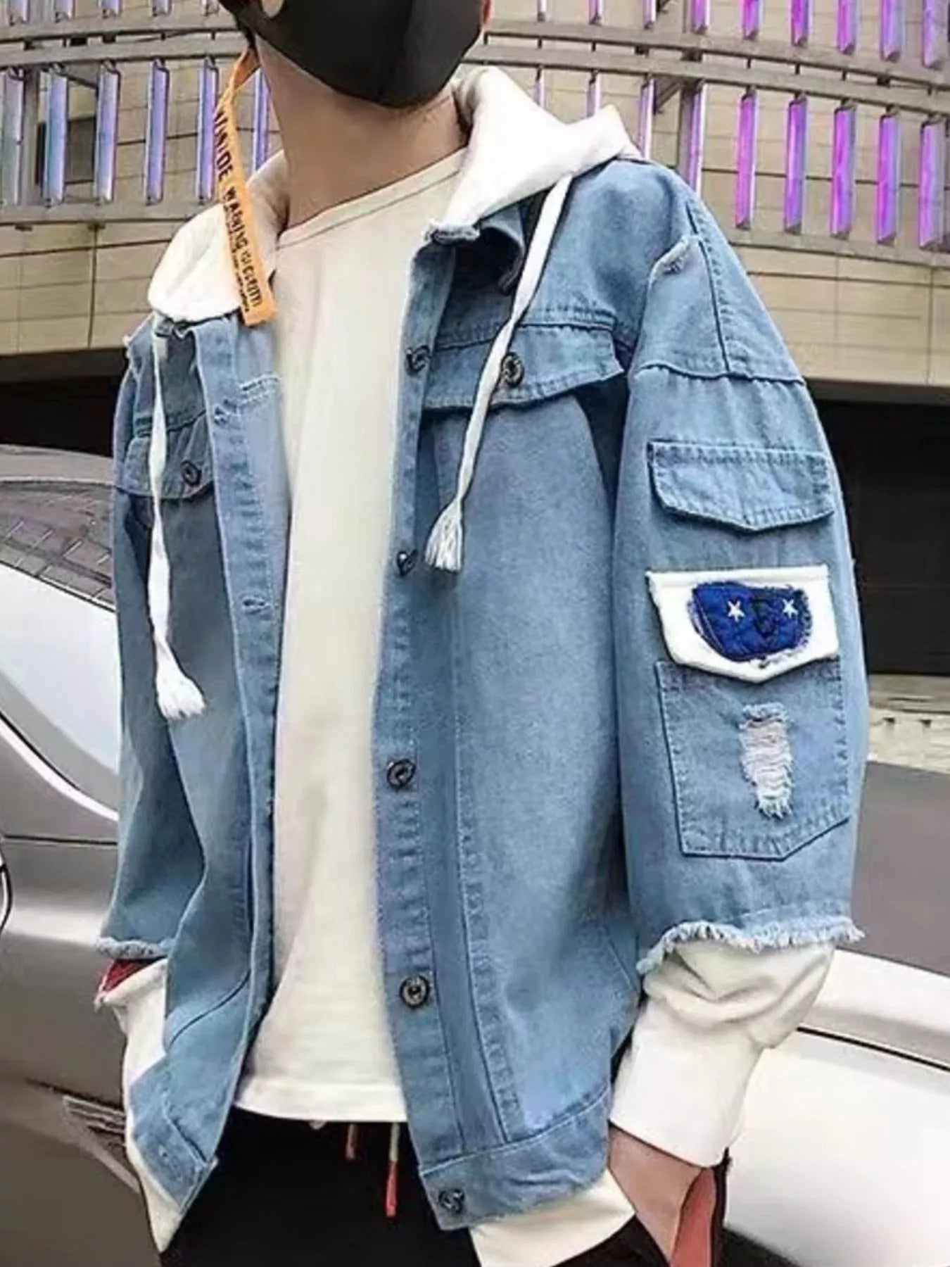 A person wearing a light blue denim jacket over a white hoodie and white t-shirt, standing outdoors near a car.