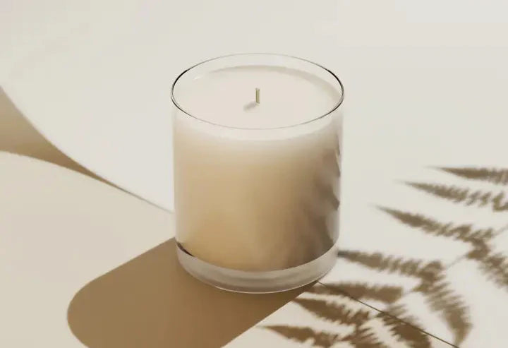 White candle in a glass container on a light surface with a leafy shadow