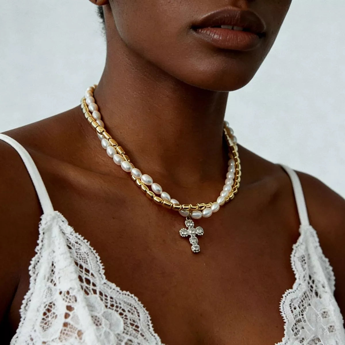 A close-up of a model wearing a double-strand necklace featuring white pearls and gold chain links with a gold cross pendant adorned with clear crystals, styled with a white lace camisole.