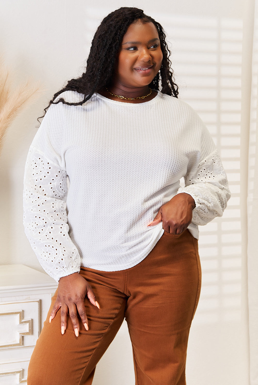 Woman wearing a light gray sweater with lace details and brown pants indoors.