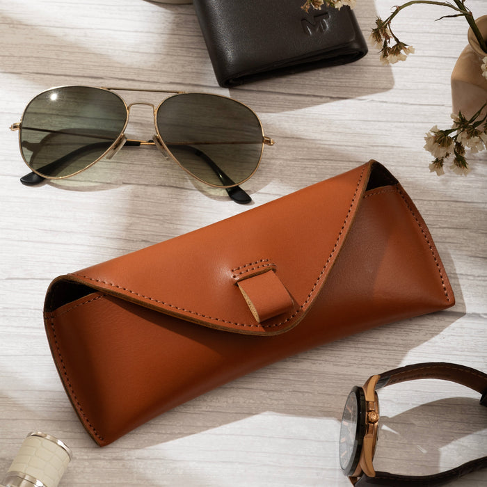Brown leather sunglasses case with sunglasses, wallet, and watch on a light wooden surface.