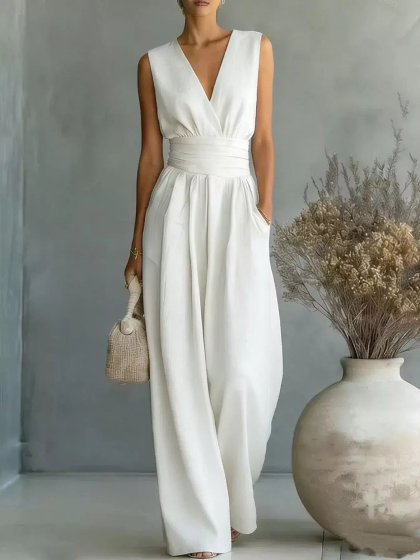 Woman wearing a white sleeveless dress standing in a room with a vase and dried plants.