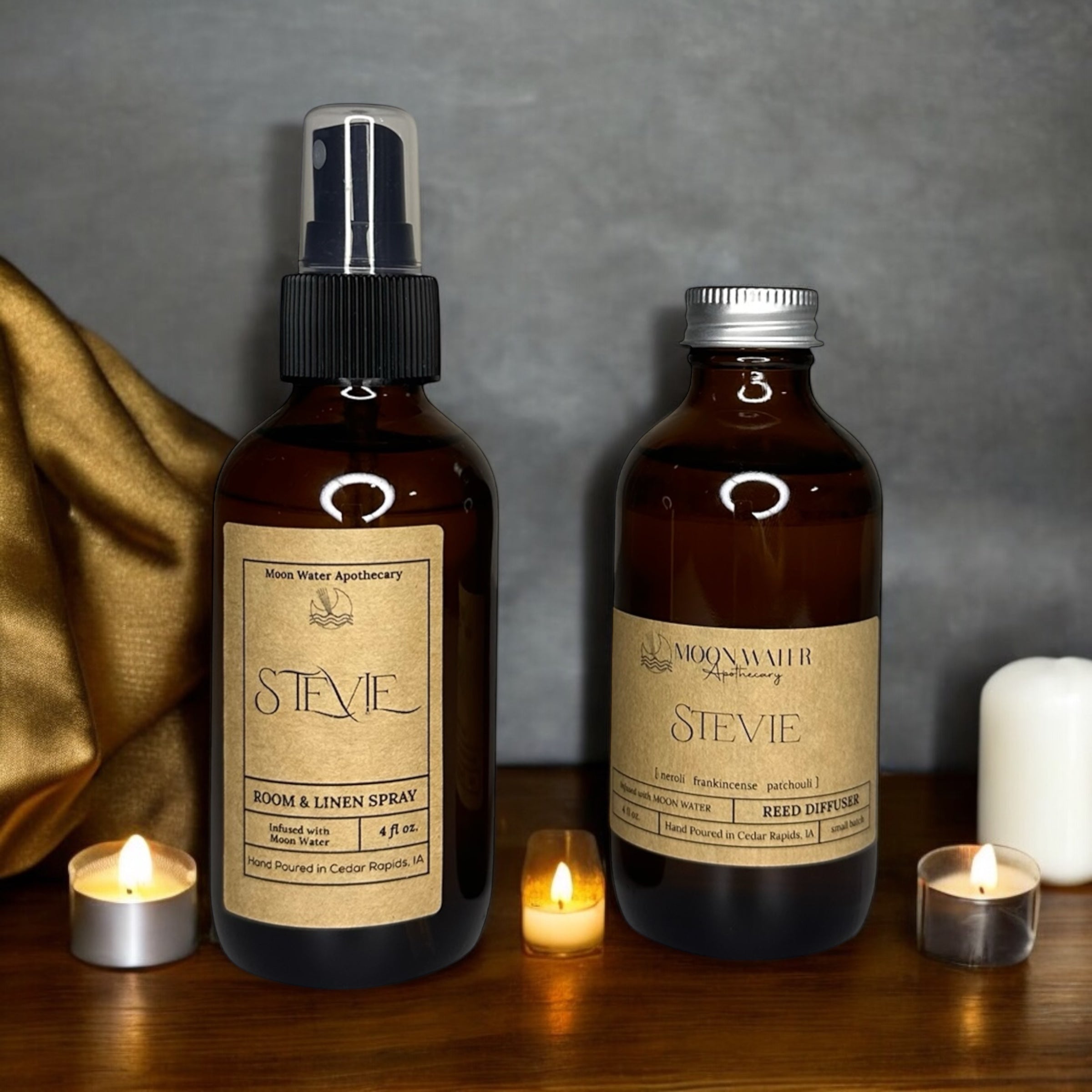 Two bottles of 'Stevie' room and linen spray and reed diffuser on a wooden surface with candles. STEVIE captures an intoxicating blend of contrasts—crisp neroli brightness meets the smoky depth of frankincense and myrrh, creating an immediately captivating opening. The fragrance's soul lies in its heart notes: a carefully balanced pairing of soft vetiver and patchouli that grounds the composition with earthy warmth and subtle complexity. As the scent develops, vanilla and amber emerge to add refinement.