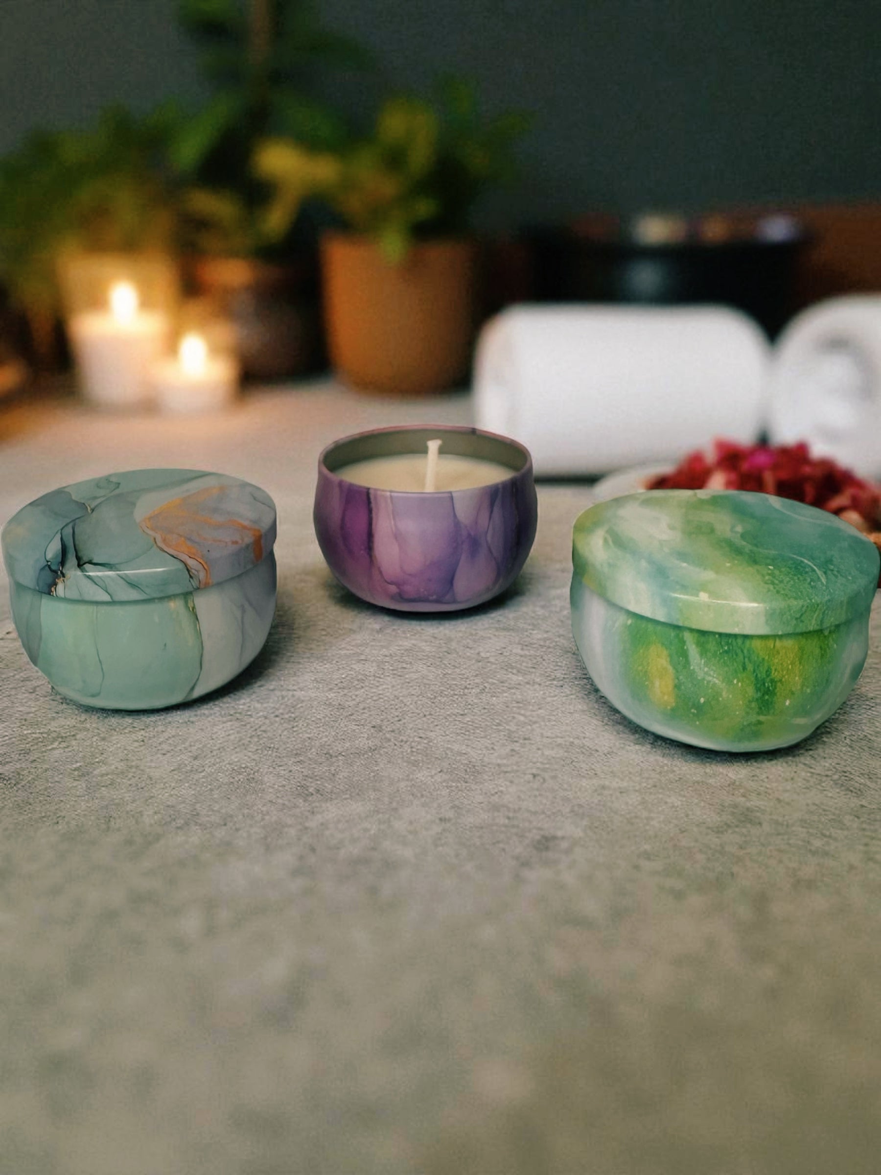 Three marbled containers with candles on a table with a blurred background. Limited-edition 2 oz tin candles in four signature scents: Zodiac (dark vetiver & sandalwood), Floral (calla lily), Marble (sea mist), and Mandala (Bulgarian lavender & sage). Once sold out, these will not be restocked.