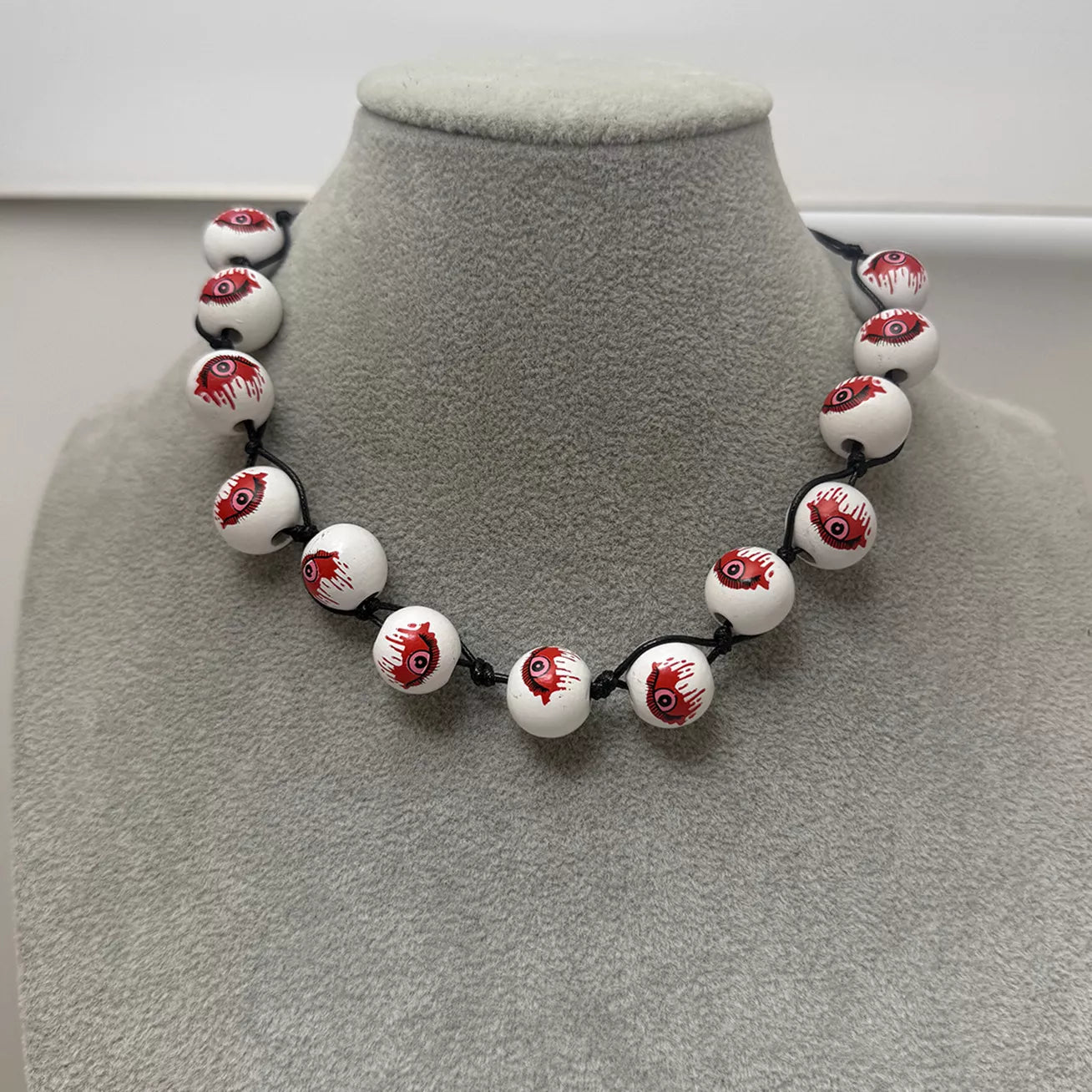 A beaded necklace displayed on a gray velvet jewelry bust. The necklace features glossy white round beads printed with a realistic red and black bloody eyeball design, strung on a black cord.