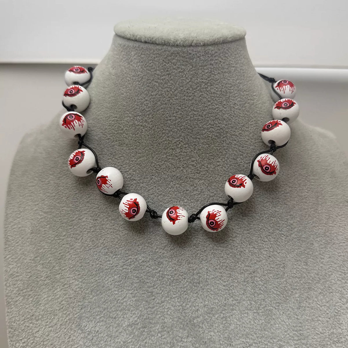 A beaded necklace displayed on a gray velvet jewelry bust. The necklace features glossy white round beads printed with a realistic red and black bloody eyeball design, strung on a black cord.