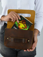 Person holding a brown leather clutch with a book inside, wearing a white shirt and gray shorts.