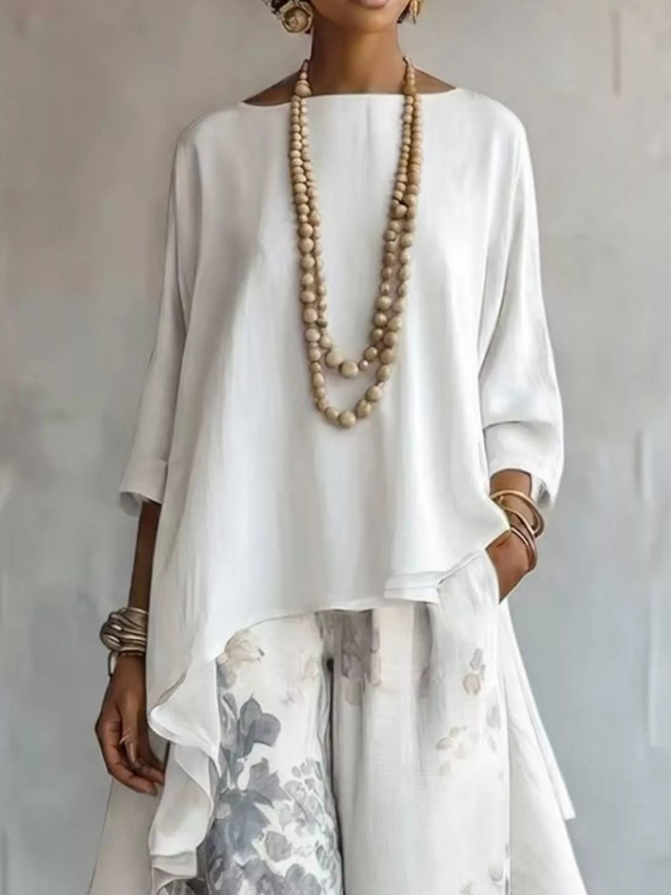 A woman wearing a loose white high-low top with three-quarter sleeves and white floral pants, accessorized with a long beaded necklace and bracelets, set against a neutral gray background.
