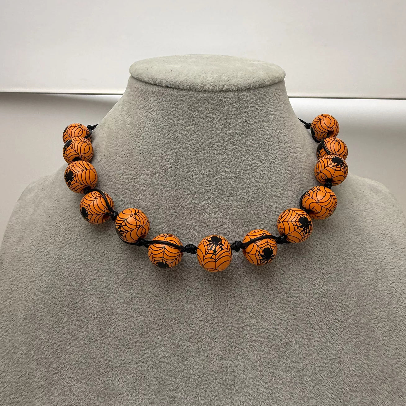An orange beaded necklace with black spiderweb and spider designs, displayed on a gray fabric jewelry bust.