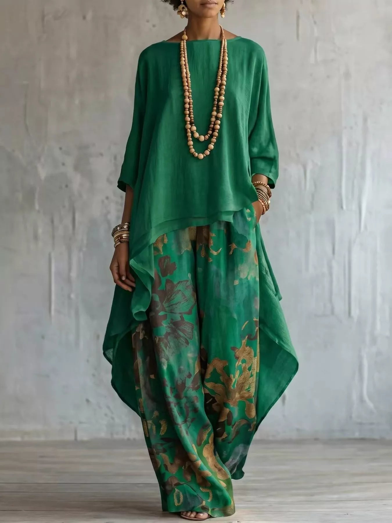 A woman models a green two-piece outfit consisting of a loose, high-low top and wide-leg pants with a floral print, accessorized with layered wooden bead necklaces and gold bracelets, set against a textured light gray wall.