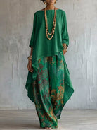 A woman models a green two-piece outfit consisting of a loose, high-low top and wide-leg pants with a floral print, accessorized with layered wooden bead necklaces and gold bracelets, set against a textured light gray wall.