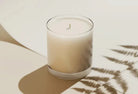 White candle in a glass container on a light surface with a leafy shadow