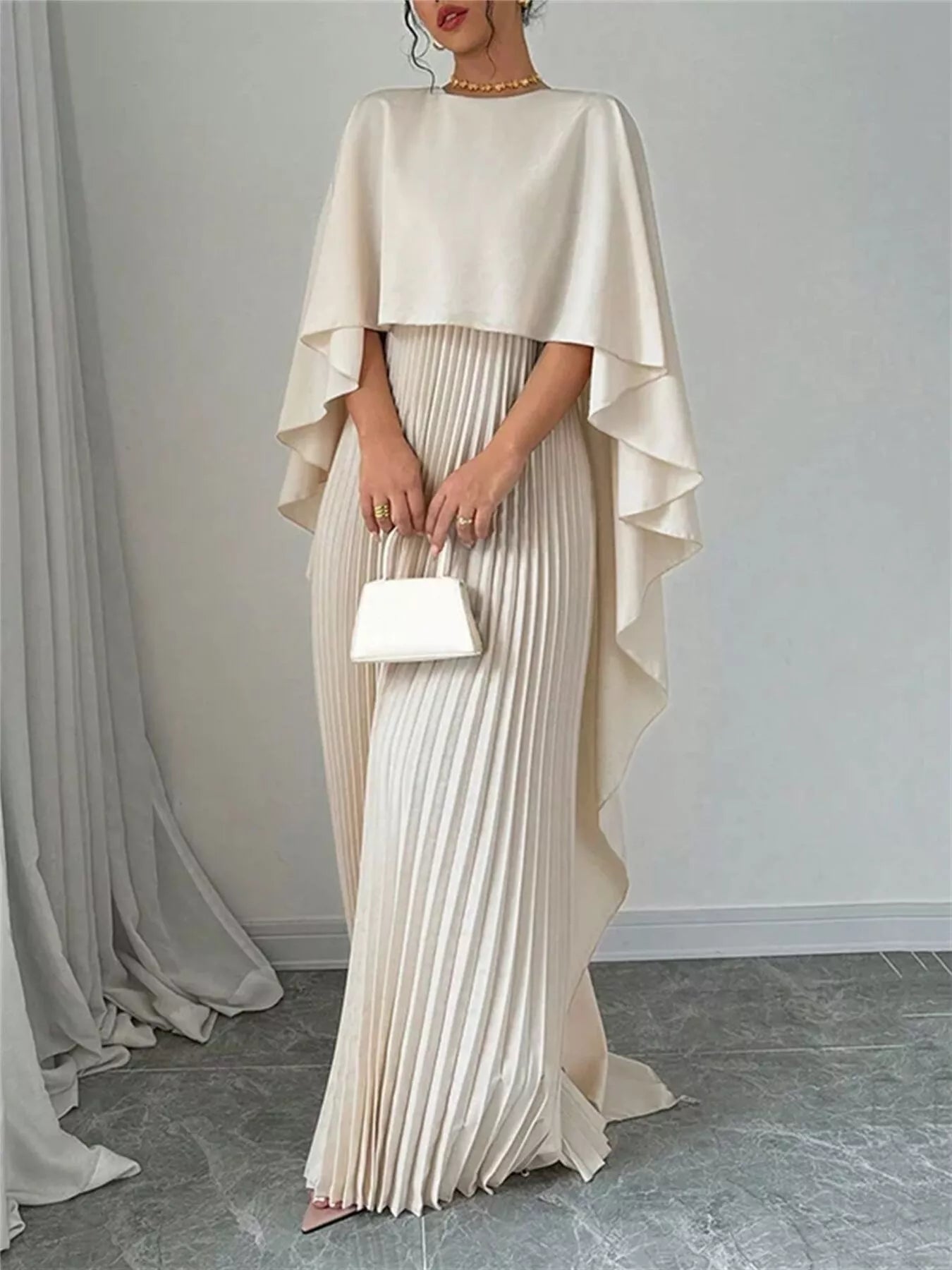 Woman wearing a beige cape over a pleated dress holding a white clutch against a plain background