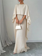 Woman wearing a beige cape over a pleated dress holding a white clutch against a plain background