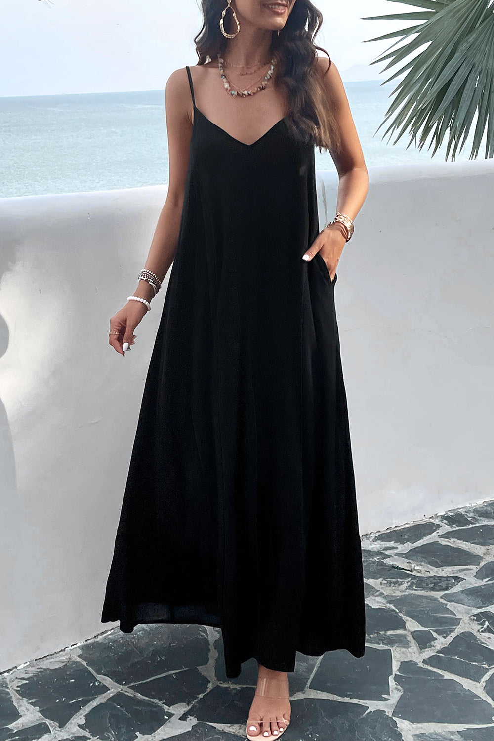 A woman wearing a black backless maxi cami dress with pockets, standing near a tropical setting.