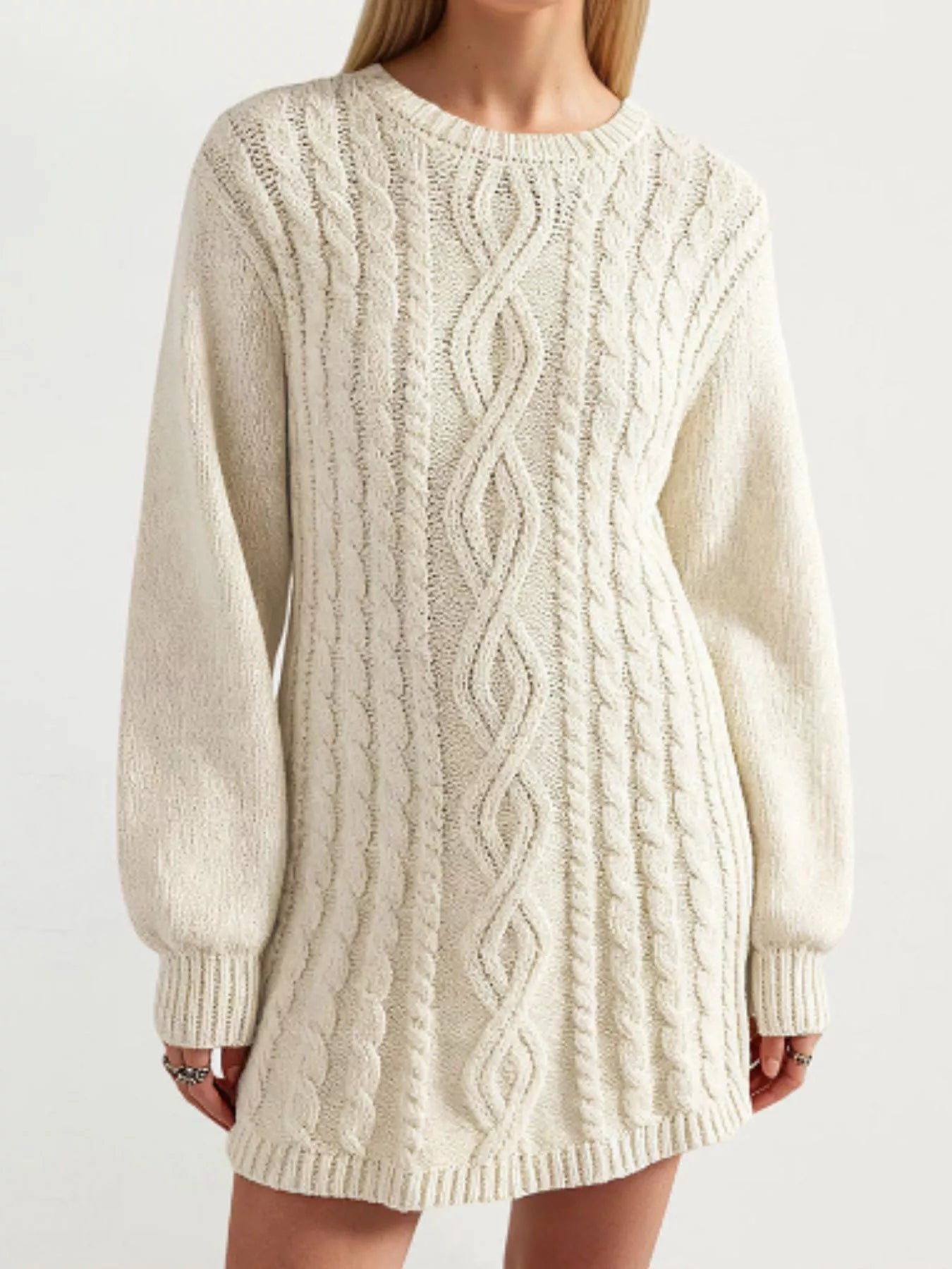 A woman wearing a cream-colored cable-knit sweater dress with a crew neck, long sleeves, and ribbed cuffs and hem, shown against a plain light background.
