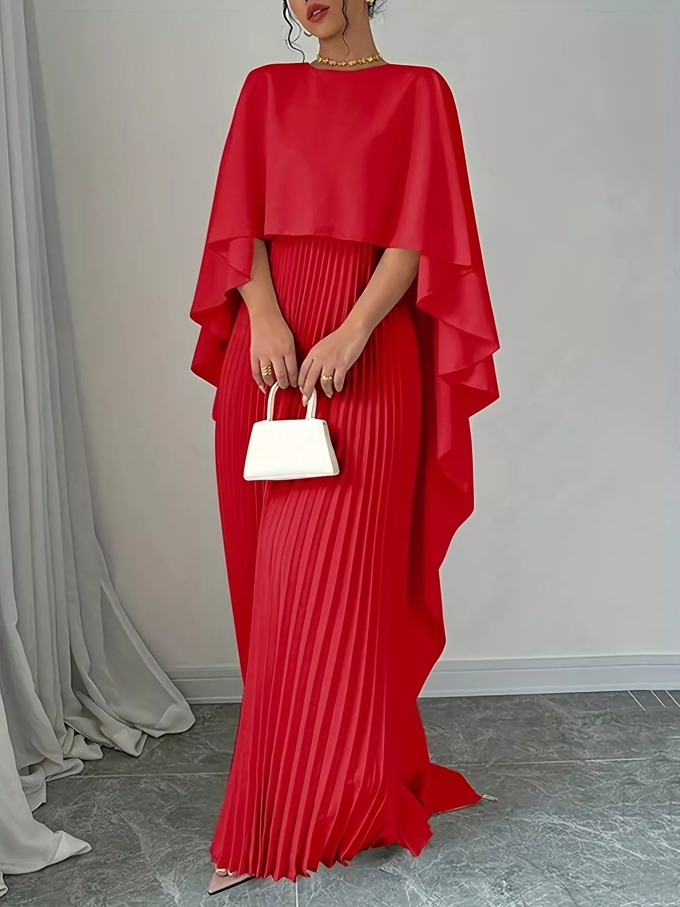 Red pleated dress with cape held by a person against a neutral background