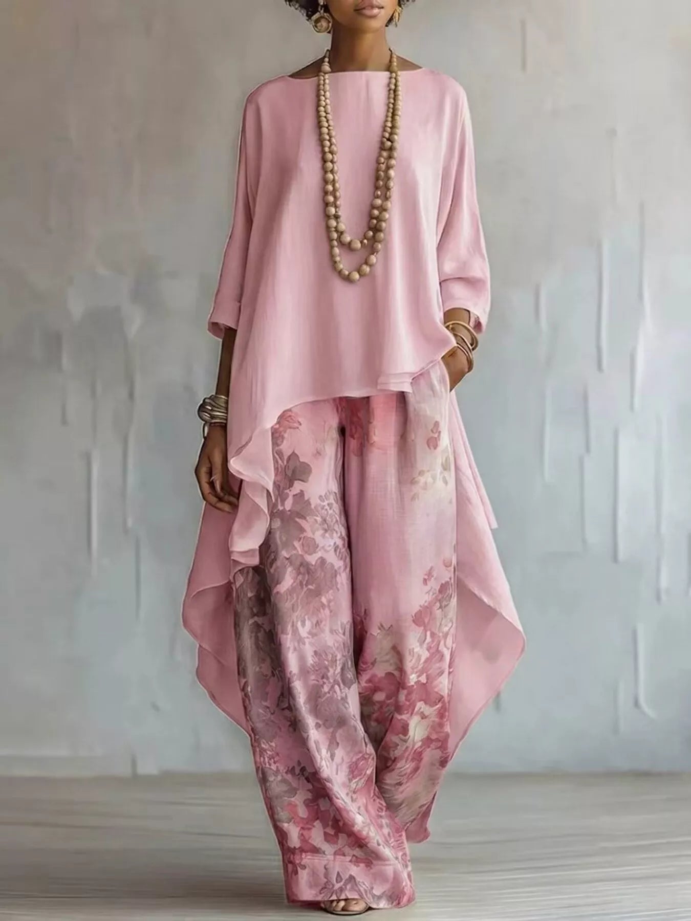 A woman models a two-piece outfit consisting of a loose, high-low pink top and matching wide-leg trousers with a pink floral watercolor print, styled with a long beaded necklace and bracelets against a textured light gray wall.