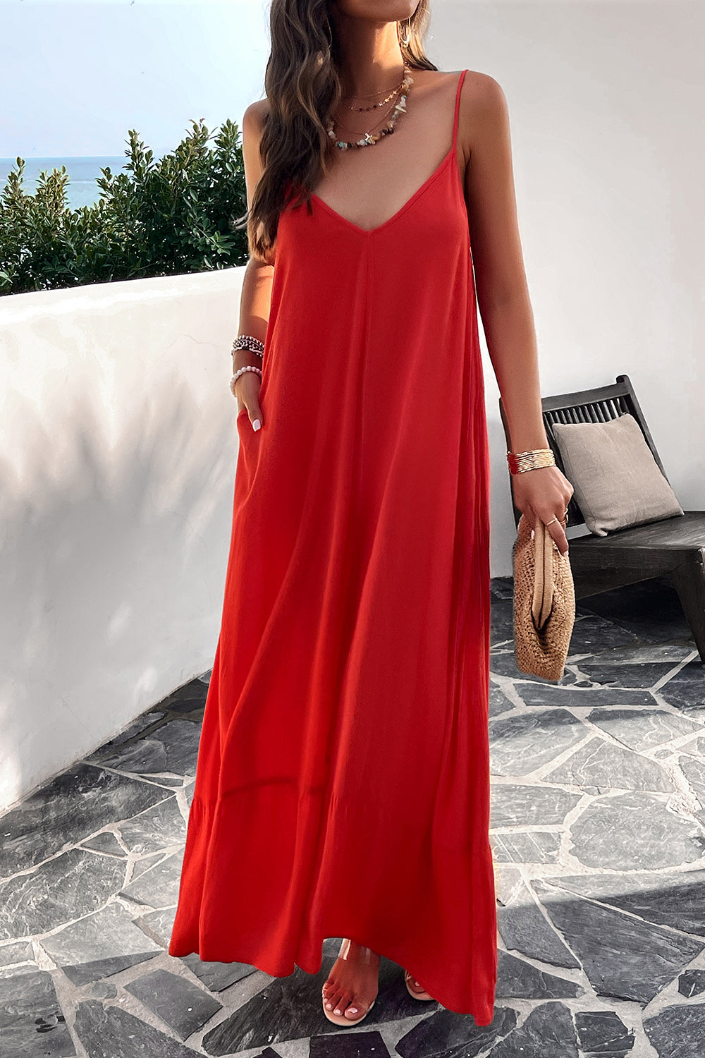 A woman wearing a red backless maxi cami dress with pockets, standing near a tropical setting.