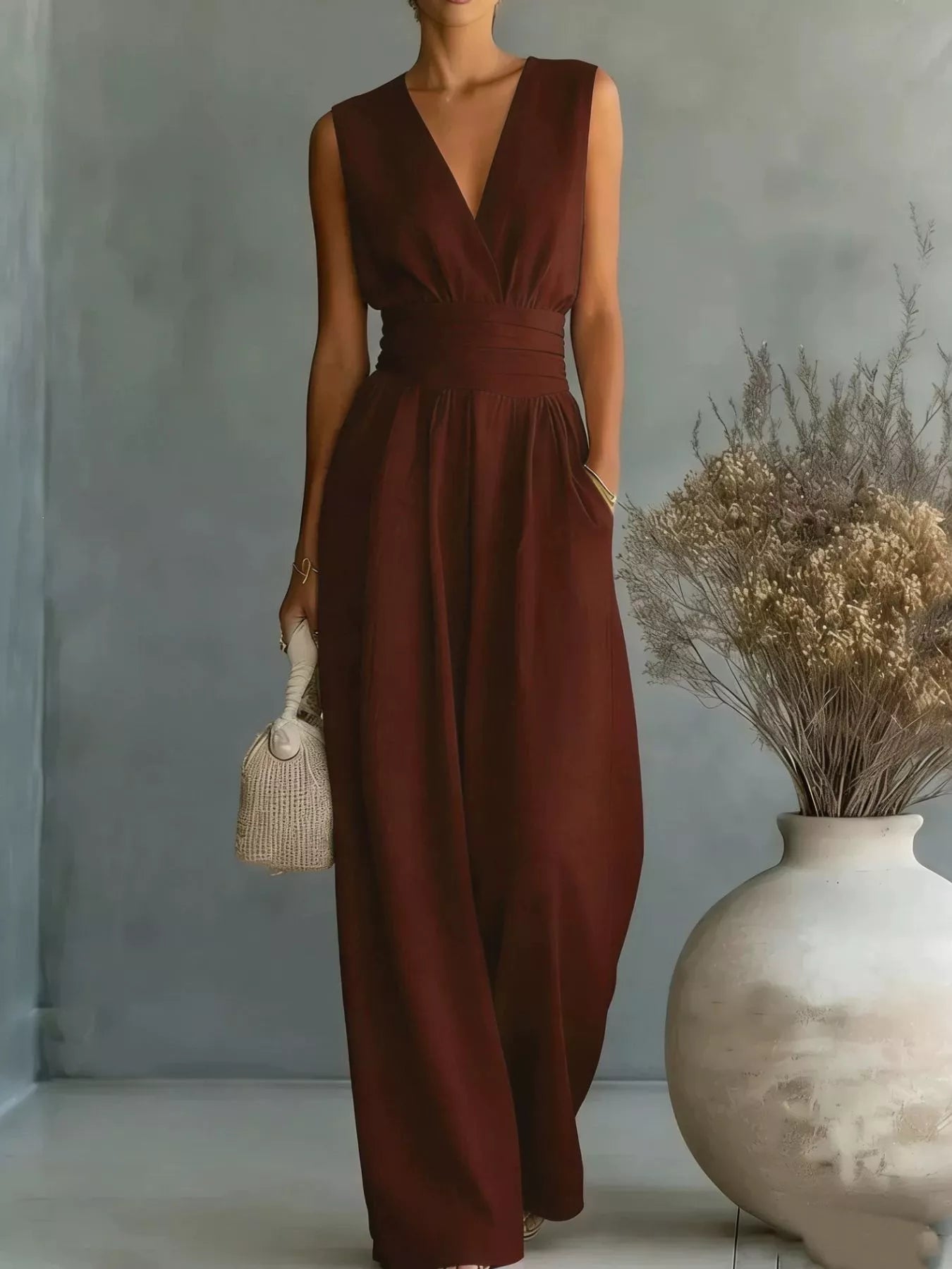 Woman wearing a brown jumpsuit holding a small bag in a minimalistic setting with a vase of dried flowers.