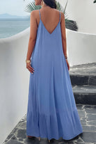 A woman wearing a blue backless maxi cami dress with pockets, standing near a tropical setting.