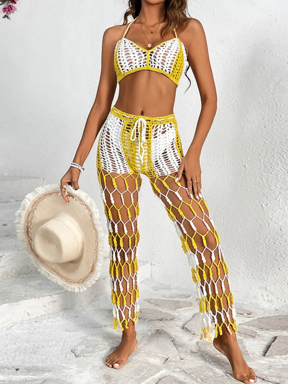 Woman wearing a yellow and white crochet two-piece swimsuit holding a hat on a light background