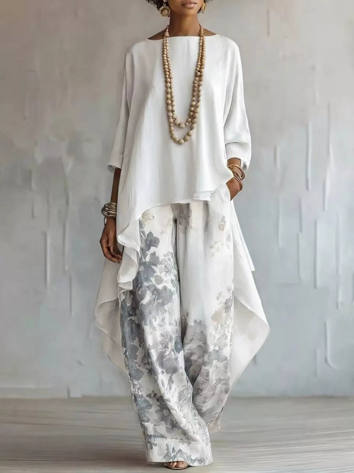 A woman models a white long-sleeve top with an asymmetrical draped hem paired with wide-leg white pants featuring a gray and blue-gray floral print. She is accessorized with layered wooden bead necklaces and bracelets, standing against a textured light gray wall.