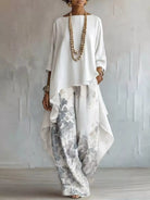 A woman models a white long-sleeve top with an asymmetrical draped hem paired with wide-leg white pants featuring a gray and blue-gray floral print. She is accessorized with layered wooden bead necklaces and bracelets, standing against a textured light gray wall.