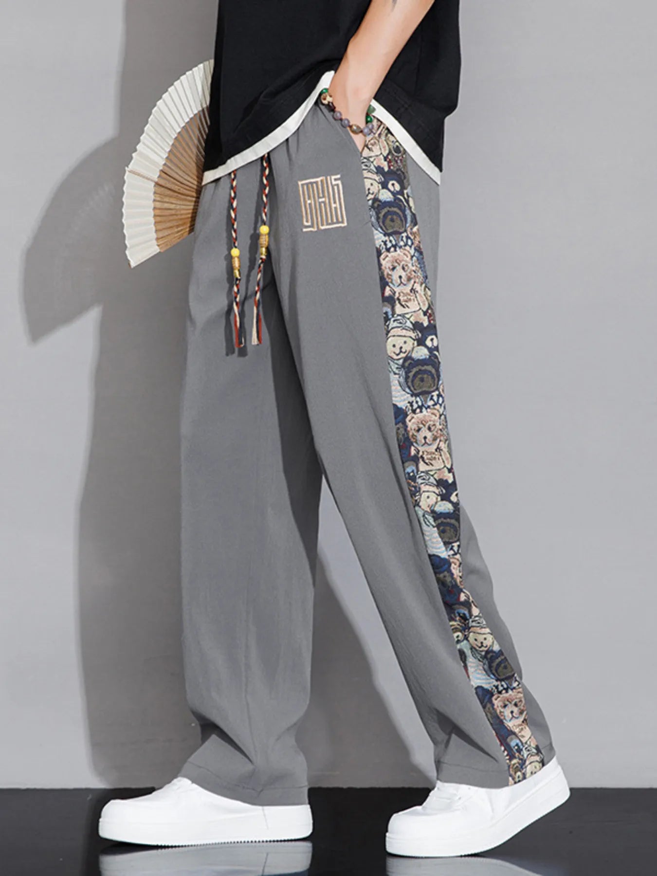 Person wearing gray pants with floral side panels, holding a fan, on a plain background