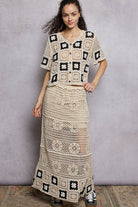 Woman wearing a crochet outfit with a patterned top and long skirt against a plain background