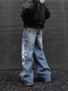 Person wearing blue jeans with a white design, standing against a dark wall.