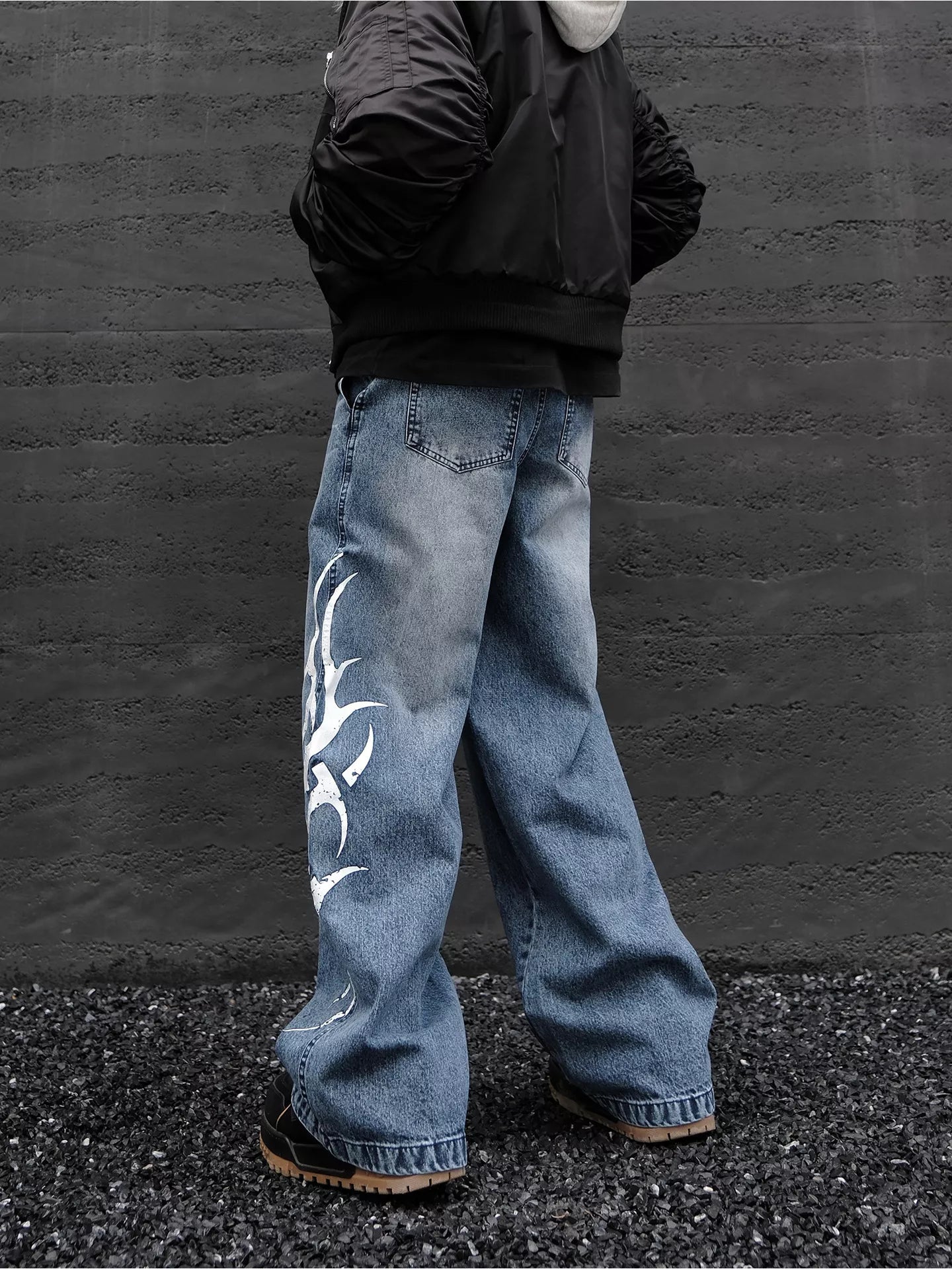 Person wearing blue jeans with a white design, standing against a dark wall.