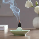 Incense cone burning on a green dish with smoke rising, placed on a wooden surface.