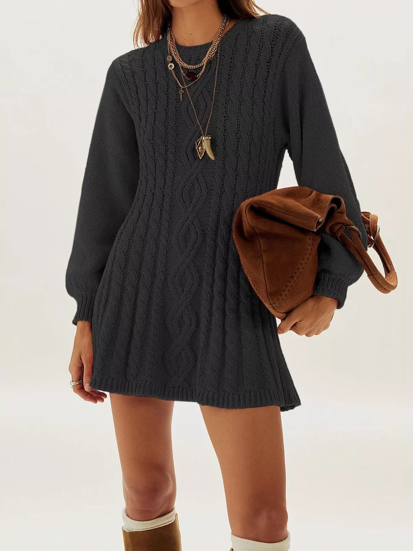 A woman wearing a dark charcoal gray cable-knit sweater dress, styled with layered gold necklaces and a brown suede shoulder bag, against a plain white studio background.