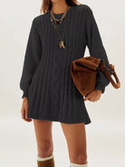A woman wearing a dark charcoal gray cable-knit sweater dress, styled with layered gold necklaces and a brown suede shoulder bag, against a plain white studio background.