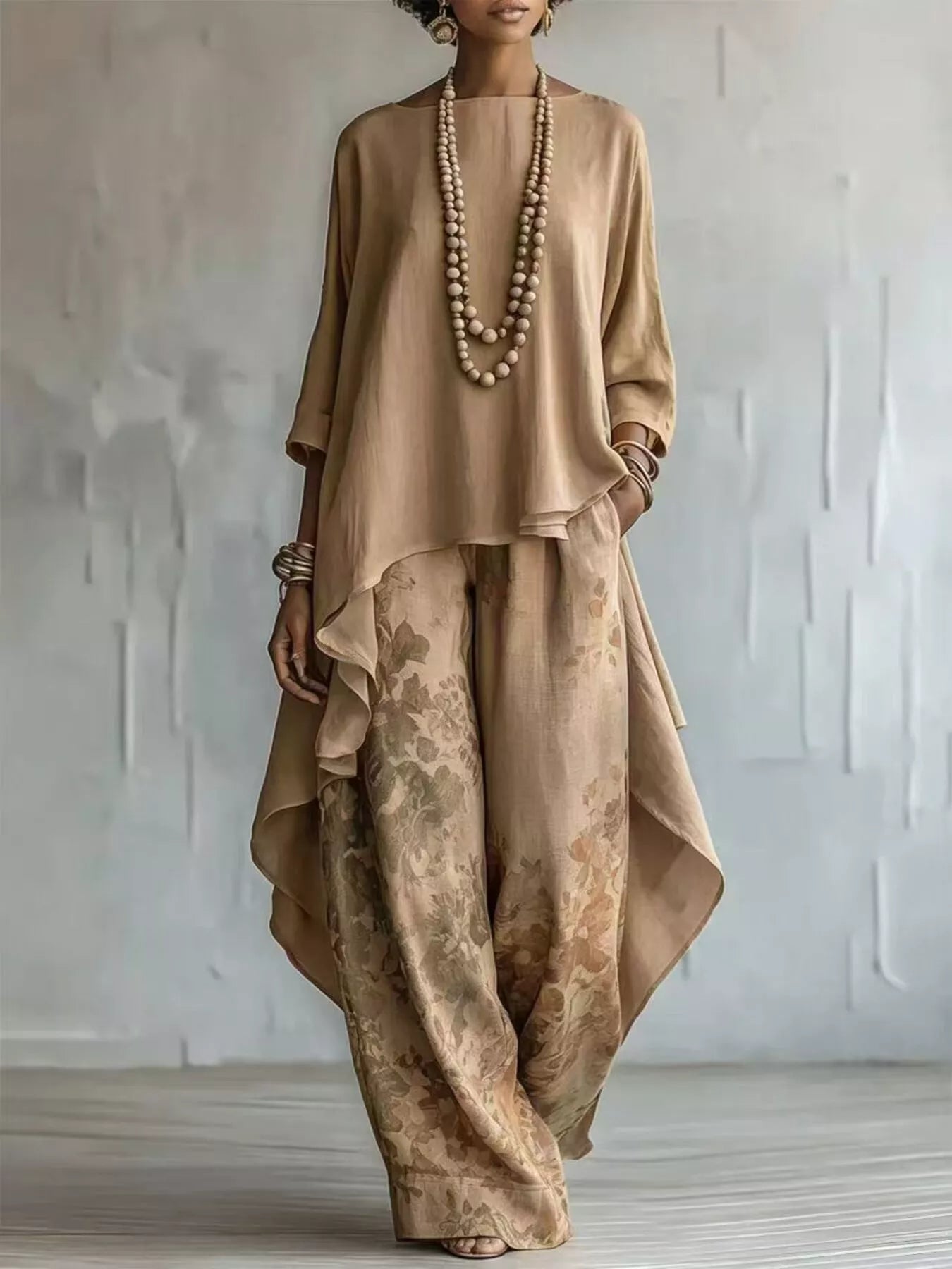 A woman models a two-piece linen outfit in a warm beige and tan floral print, featuring a loose, asymmetrical long-sleeve top and wide-leg pants, accessorized with a long beaded necklace and bracelets against a textured light gray wall.