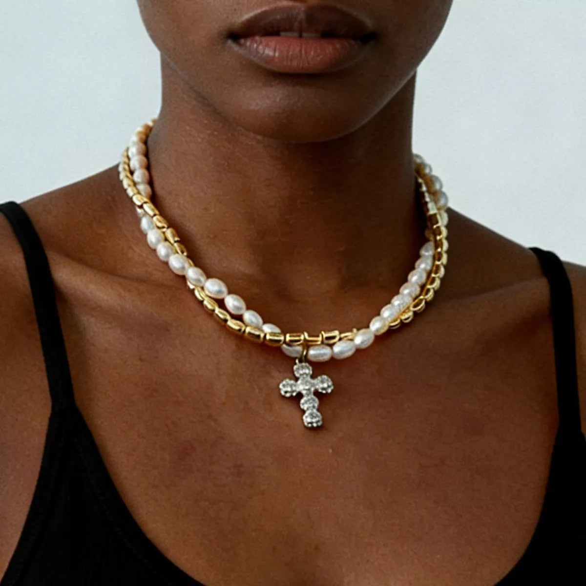 A close-up of a model wearing a double-strand necklace featuring white pearls and gold-toned chain links, with a silver-toned cross pendant adorned with small clear stones.