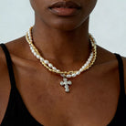 A close-up of a model wearing a double-strand necklace featuring white pearls and gold-toned chain links, with a silver-toned cross pendant adorned with small clear stones.