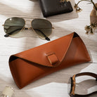 Brown leather sunglasses case with sunglasses, wallet, and watch on a light wooden surface.
