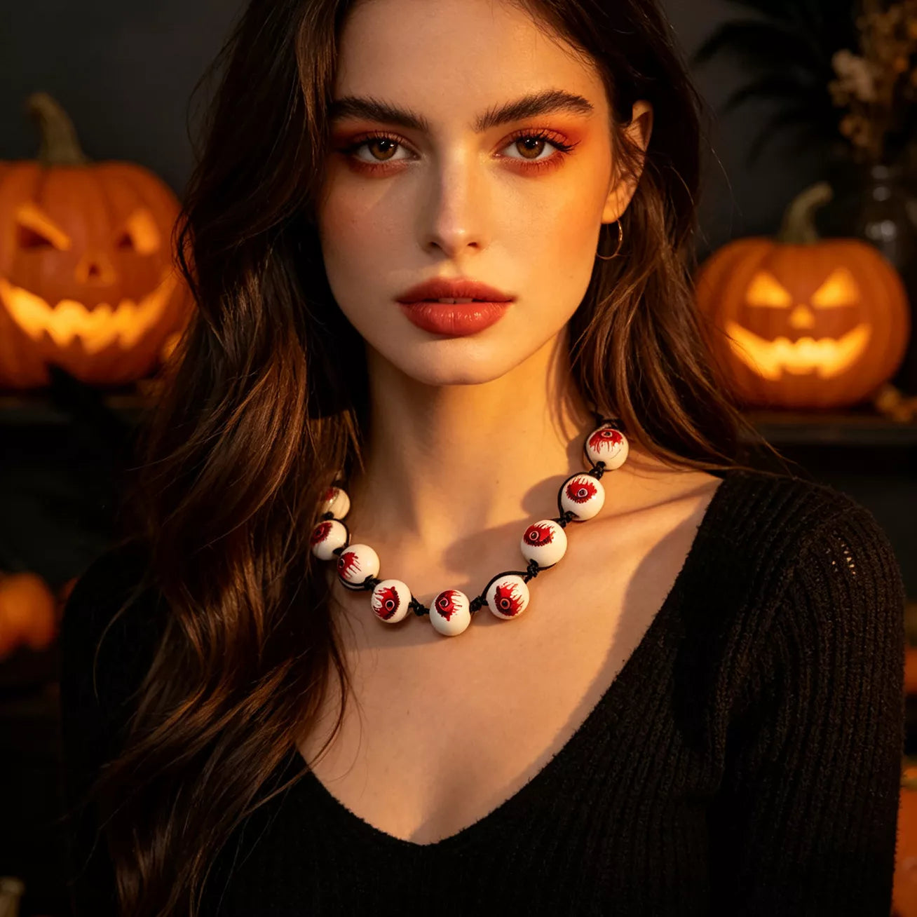 A woman with long dark hair and warm-toned makeup wears a chunky beaded necklace made of white beads with red and black details, posed in front of glowing jack-o'-lanterns.