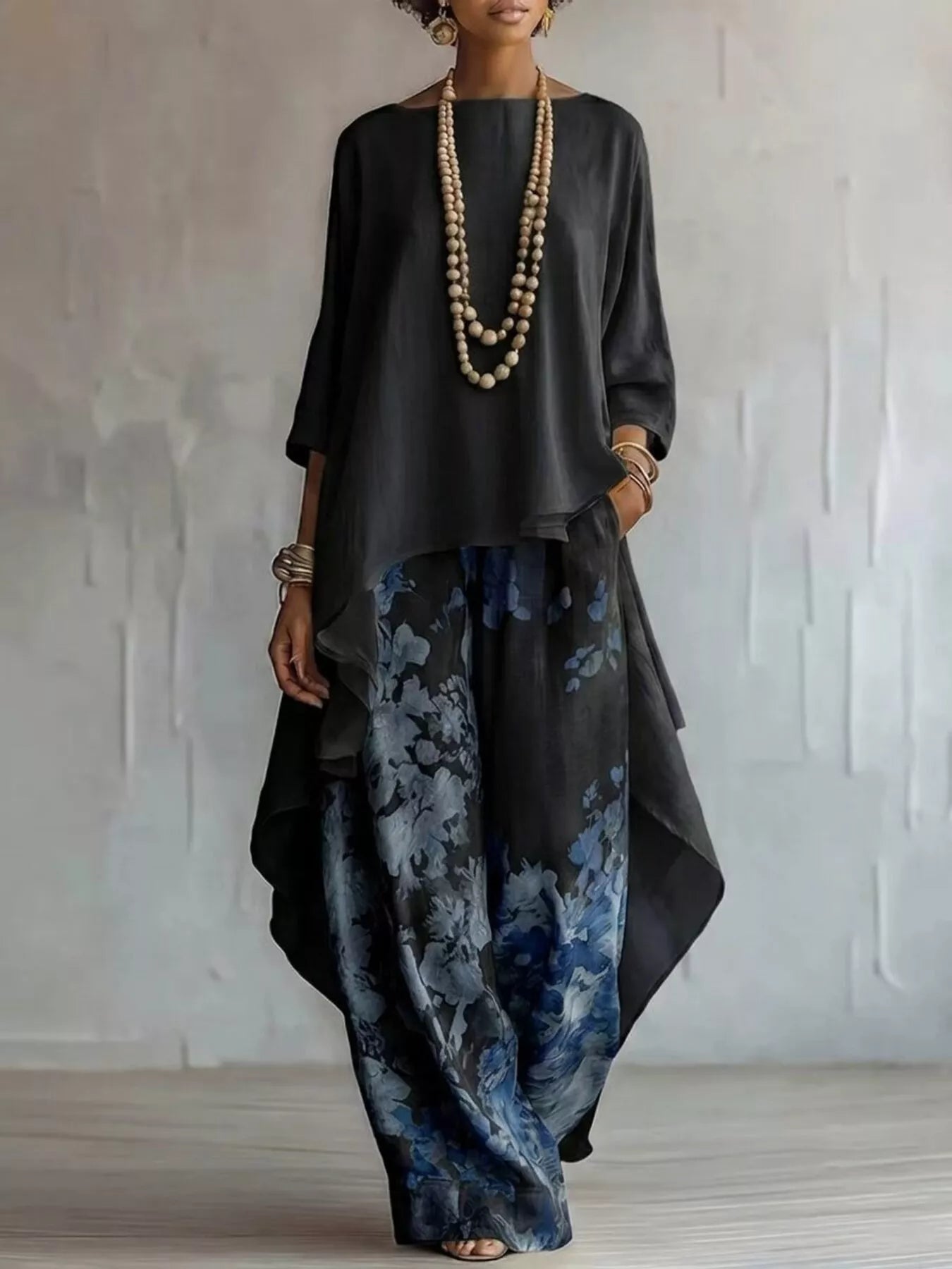 A woman models a loose-fitting black asymmetrical top paired with wide-leg blue and gray floral print pants, accessorized with a long beaded necklace and bracelets, set against a textured light gray wall.
