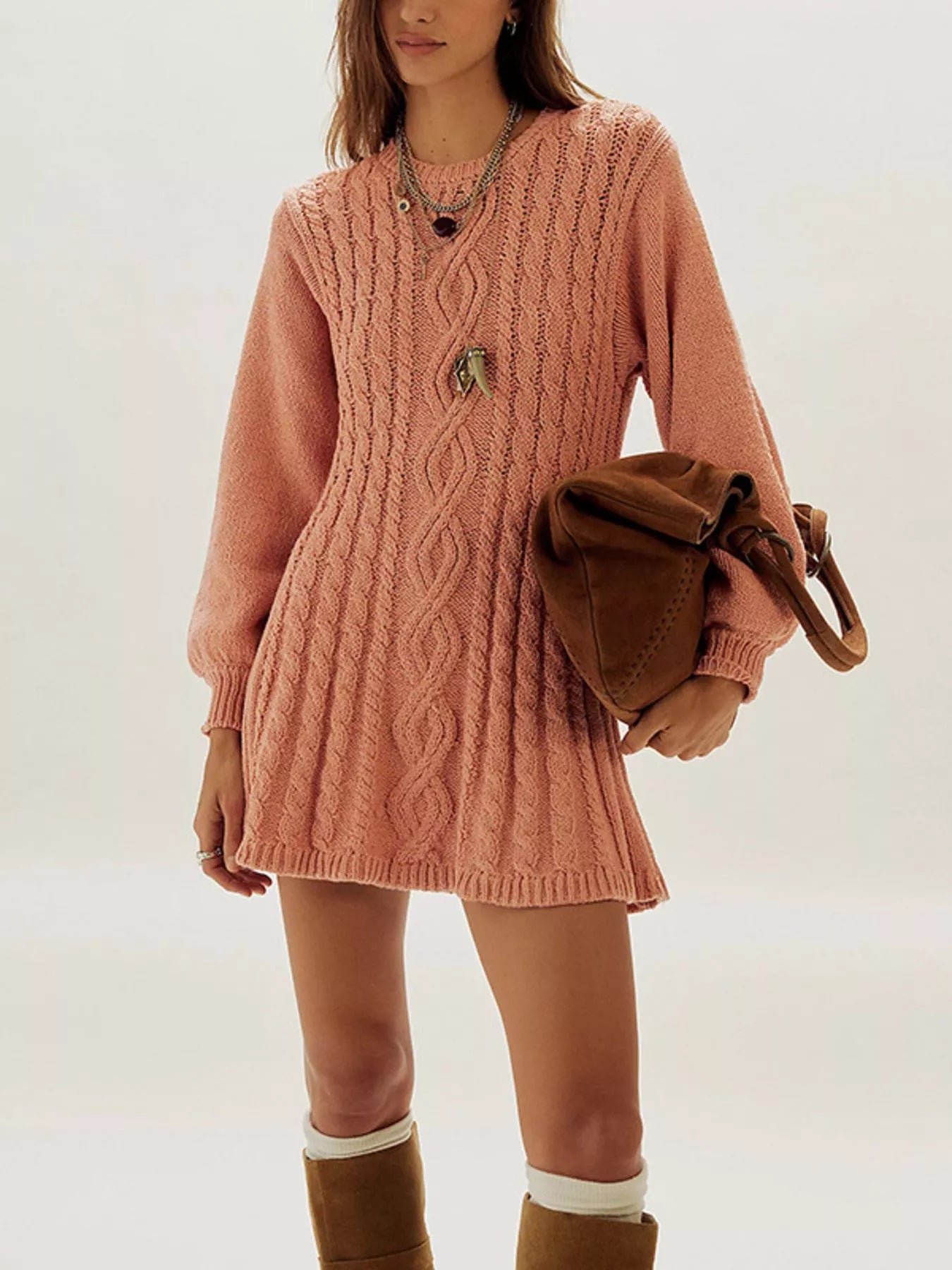 A woman models a dusty pink cable-knit sweater dress with long sleeves and a crew neck, styled with a brown leather shoulder bag and brown boots against a plain light background.