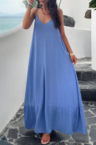 A woman wearing a blue backless maxi cami dress with pockets, standing near a tropical setting.