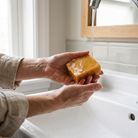 Hands holding a turmeric soap bar under running water over a white bathroom sink.