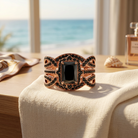 Rose-gold-tone wedding ring set with rectangular black CZ and infinity-style bands on ivory linen beside an ocean-view window.